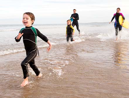 Family of four are running along the shore with body boards. They are all laughing.