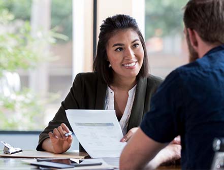 A cheerful female banker sits at her desk while speaking to a client about easy account transfers. 