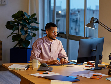 A man sits with glasses sits at his work from home desk in the evening, focusing on the computer screen. 
