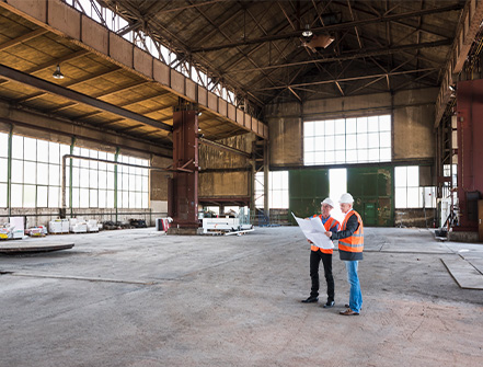 两名身穿橙色背心、头戴白色头盔的男子站在一个大仓库内，举着他们下一个商业项目的建筑文件。