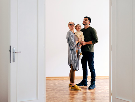 Una mamá y un papá jóvenes sostienen a un niño en sus brazos mientras miran una casa nueva que está a la venta. 