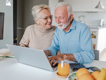 Una pareja de mediana edad se sienta en la mesa de la cocina mientras se sonríen y miran sus finanzas de administración de patrimonio en una computadora.