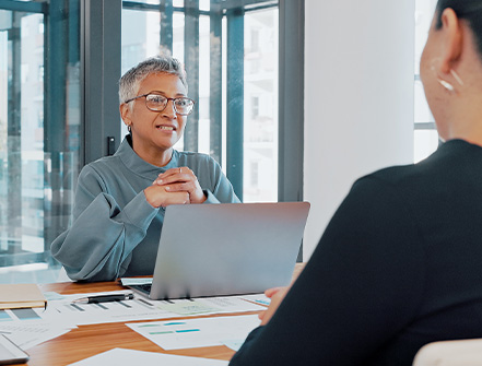 A business client sits while facing an auditing advisor inside a bright office while they review paperwork over a wooden desk.