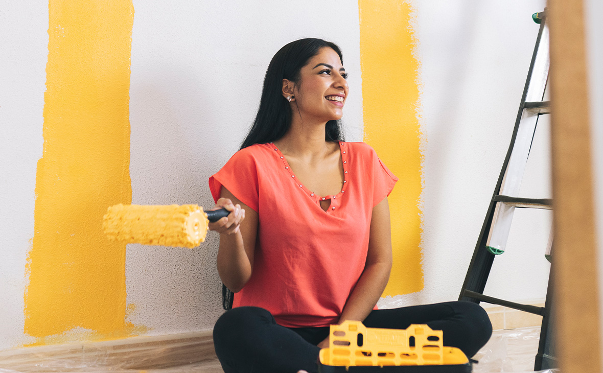 Una joven sonriente con un rodillo de pintura sentada junto a una escalera en la casa