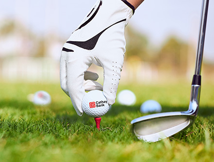 A Cathay Bank Golf Ball sits on the grass at an outdoor golf course with a golf club behind it.
