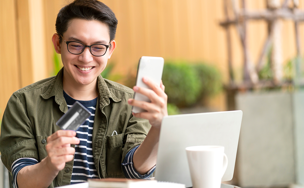A man is sitting at a table outdoors with a laptop, holding a smartphone in one hand and a credit card in the other, suggesting an online payment or mobile banking activity.