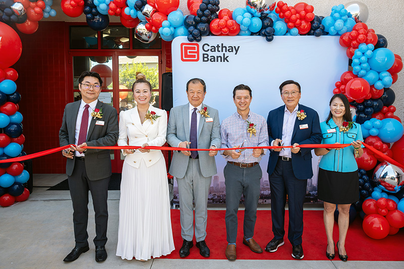 Six people stand on a red carpet outside a Cathay Bank location, holding scissors and cutting a red ribbon during a ribbon-cutting ceremony. A large balloon arch in red, light blue, navy, and silver frames the entrance and a backdrop displaying the Cathay Bank logo