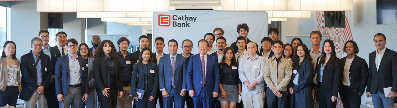 A large group of students and Cathay Bank representatives standing together in front of a Cathay Bank-branded backdrop. The group is dressed in business or business casual attire, posed for a formal group photo inside the corporate offices, with modern lighting and interior décor in the background.