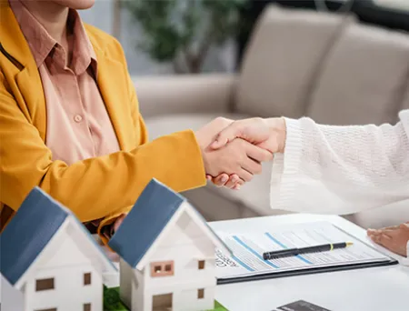 Two individuals sit at a table and shake hands over a mortgage refinancing agreement, with miniature house models, a clipboard of loan documents, and a pen on the table, symbolizing a completed home refinance deal.