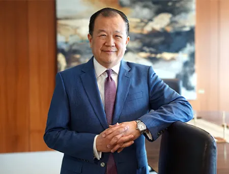 Cathay Bank's President and CEO, Chang M. Liu, in a blue business suit with a white shirt and tie stands in a modern office, hands clasped and resting on a chair, with a wood-paneled wall and abstract painting in the background.
