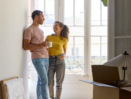 Two adults stand together in a bright apartment near tall windows, surrounded by moving boxes and a table lamp, suggesting they are settling into a new home.