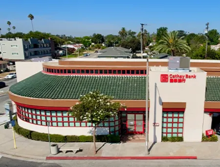Exterior view of Cathay Bank’s Alhambra Branch, featuring a cream-colored building with a green tile roof, red-framed windows, and prominent Cathay Bank signage. 