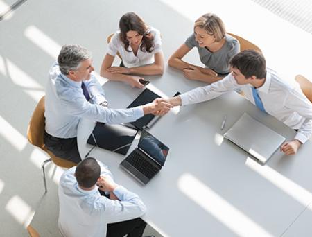 A group of professionals are gathered around a conference table shaking hands as they make a business deal. 