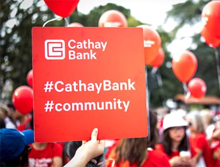 A red sign is being held by a person in a crowd which shows the Cathay Bank logo as well as hashtags for community and Cathay Bank