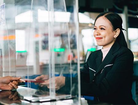 A bank teller stands behind a protective glass shield while handing a checkbook back to a client.