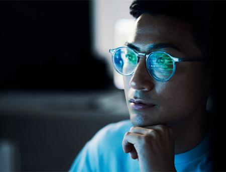 Hay un primer plano del rostro de un joven con lentes, que refleja su actividad en pantalla desde una computadora.