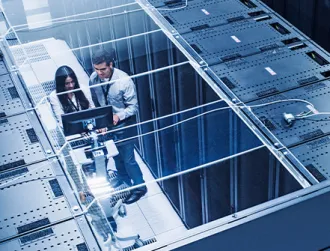 Two tech industry professionals stand in between a row of server racks while analyzing data on a computer.