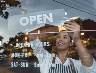 A small business owner adjusts an “open” sign on the front window of a shop. Reflections of the street, cars, and warm interior lights show a lively neighborhood environment.