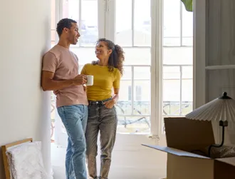 Two adults stand together in a bright apartment near tall windows, surrounded by moving boxes and a table lamp, suggesting they are settling into a new home.