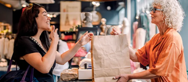 Client and retail worker exchange a shopping bag across a boutique clothing store counter, with racks of apparel and warm lighting in the background.