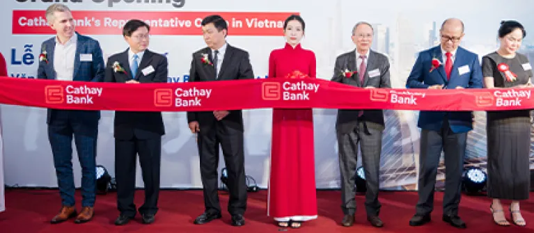 Ribbon-cutting ceremony at the grand opening of Cathay Bank's Vietnam Representative Office, with attendees holding a branded red ribbon before a Cathay Bank backdrop.