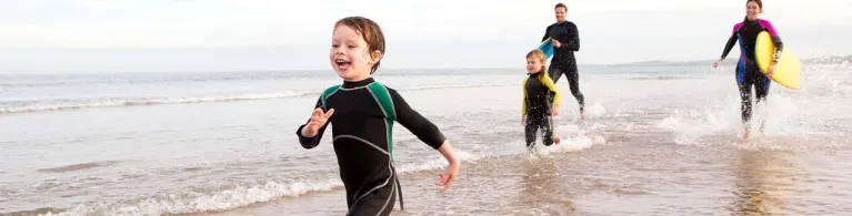 Family of four are running along the shore with body boards. They are all laughing.