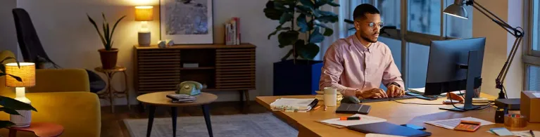 A man sits with glasses sits at his work from home desk in the evening, focusing on the computer screen. 