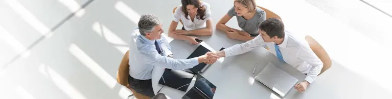 A group of professionals are gathered around a conference table shaking hands as they make a business deal. 