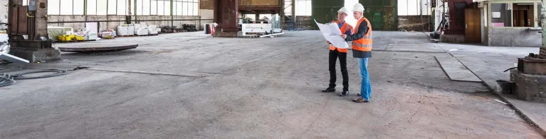 Two men in orange vests and white helmets stand inside a large warehouse holding up construction papers for their next business venture
