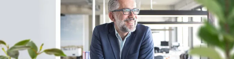 A middle-aged man with glasses and grey hair smiles as he leans over a window inside his small business office. 