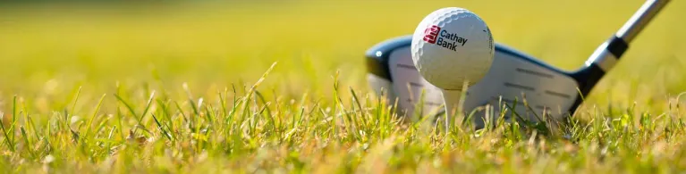 A Cathay Bank Golf Ball sits on the grass at an outdoor golf course with a golf club behind it.