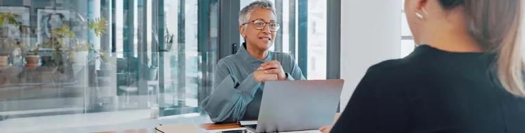 A business client sits while facing an auditing advisor inside a bright office while they review paperwork over a wooden desk.