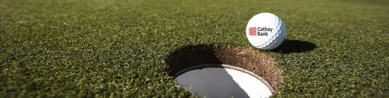 A Cathay Bank Golf Ball sits on the grass at an outdoor golf course with a golf club behind it.