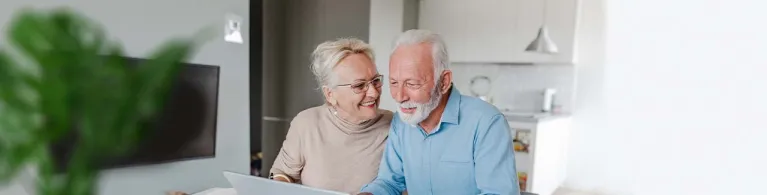 A middle-aged couple sits at a kitchen table while smiling at each other and looking at their wealth management finances on a laptop.