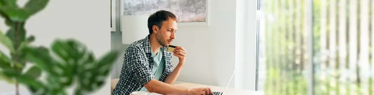 A man sits at his wooden desk in a brightly lit office while staring at his laptop and holding his credit card with a worried look about fraud alerts