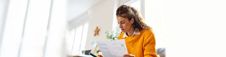 A woman wearing a yellow sweater looks cozy while sitting on the couch in her living room while holding important financial documents and checking her credit history on her cell phone.