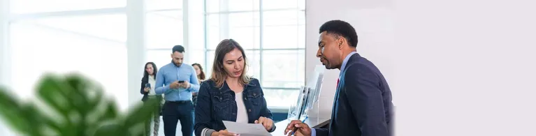 An adult male bank manager stands at the counter to answer the adult woman's questions about her monthly statements and recent check cashing.