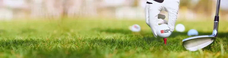A Cathay Bank Golf Ball sits on the grass at an outdoor golf course with a golf club behind it.