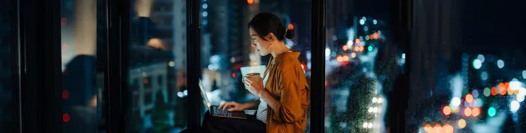 A woman in an orange shirt sits in front of her laptop in a dimly lit apartment, checking to report a phishing scam online.