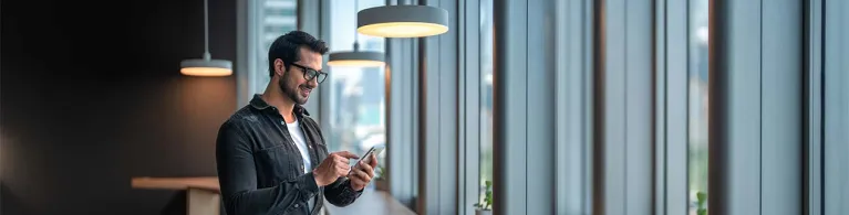 A male wearing glasses in a well-lit office space with warm lamps hanging from the ceiling stares happily at his phone while receiving security protection alerts from his mobile banking app.