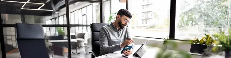 A man in a grey quarter-zip sweater and glasses sits at his work desk. In his left hand he is holding a phone, while his right hand is focused and pointing toward an iPad lying on the desk in front of him. 