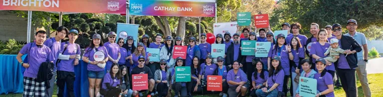 Cathay Bank team members in purple t-shirts stand together in an outdoor grass area with a Cathay Bank tent behind them, holding Cathay Bank signs for a group photo.