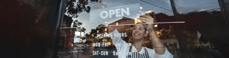 A small business owner adjusts an “open” sign on the front window of a shop. Reflections of the street, cars, and warm interior lights show a lively neighborhood environment.