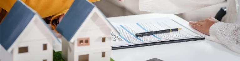 Two individuals sit at a table and shake hands over a mortgage refinancing agreement, with miniature house models, a clipboard of loan documents, and a pen on the table, symbolizing a completed home refinance deal.