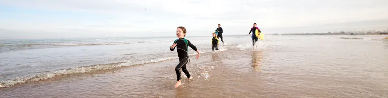 Family of four are running along the shore with body boards. They are all laughing.