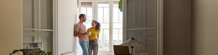 Two adults stand together in a bright apartment near tall windows, surrounded by moving boxes and a table lamp, suggesting they are settling into a new home.