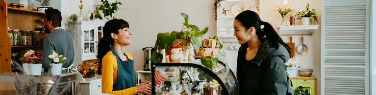 Small business owner and their shop customer converse at a café counter with a glass pastry display, surrounded by plants and warm lighting.