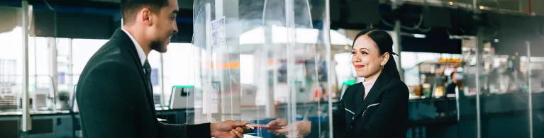 A bank teller stands behind a protective glass shield while handing a checkbook back to a client.