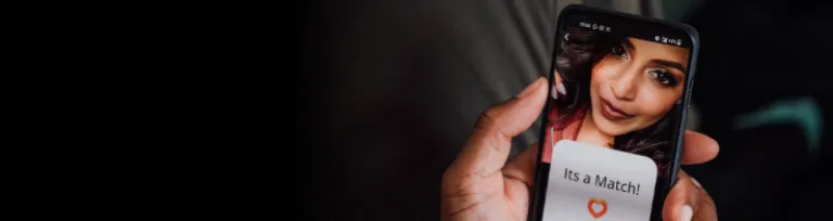 A man holds his smartphone between his hands and views a potential romantic match on a dating app which depicts the image of a young female.