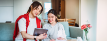 Two women sit on a couch, smiling as they review information on a tablet and a printed document in a bright living room.